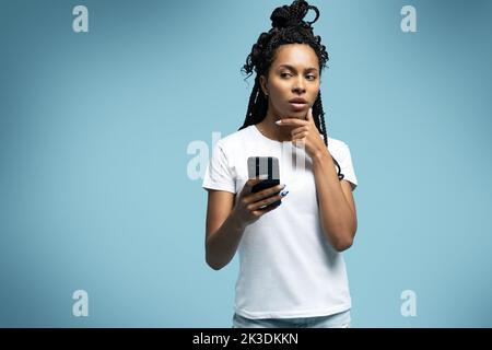 Une femme ethnique aux cheveux mauriques positifs utilise un téléphone portable vérifie les messages et lit les nouvelles tient le cellulaire moderne dans les mains regarde avec curieux heureux expression Banque D'Images