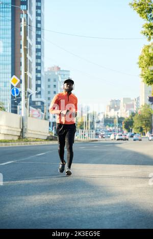 Vue de face d'un jeune athlète afro-américain en veste de sport rouge, leggiins noirs, baskets et casquette de baseball sur la route asphaltée Banque D'Images
