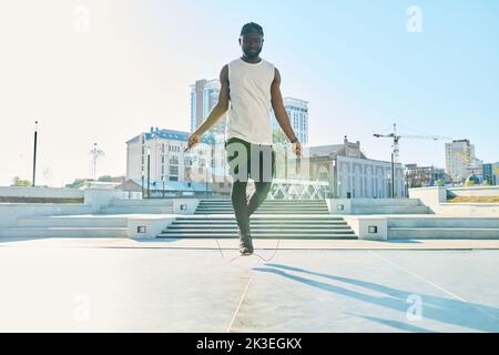 Jeune Afro-américain actif dans les vêtements de sport sautant sur la corde à sauter tout en s'exerçant sur la grande place ensoleillée le matin Banque D'Images