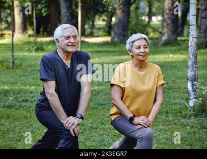 Les conjoints âgés font des exercices sportifs à l'extérieur dans le parc d'été le matin. Un couple plus âgé en bonne santé, en train de se réchauffer, en faisant des squats sourire, profitez d'une activité active Banque D'Images