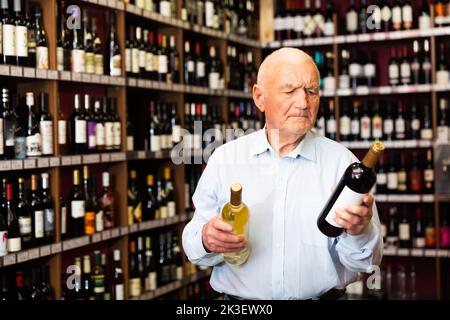 Un homme âgé sérieux choisit entre le vin rouge et le vin blanc dans un magasin de spiritueux Banque D'Images