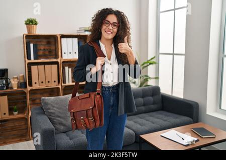 Jeune femme hispanique au bureau de consultation souriant heureux et positif, pouce vers le haut faisant excellent et signe d'approbation Banque D'Images