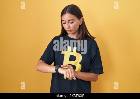 Jeune femme hispanique portant un t-shirt bitcoin vérifiant l'heure sur la montre de poignet, détendue et confiant Banque D'Images