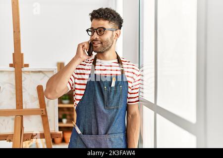 Jeune artiste arabe homme souriant heureux en utilisant un smartphone dans un studio d'art. Banque D'Images