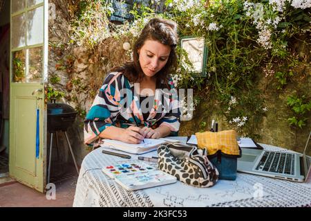 femme adulte caucasienne française assise sur une table avec un carnet et aquarelle sur la terrasse de sa maison avec un mur avec des nénuphars à l'arrière Banque D'Images