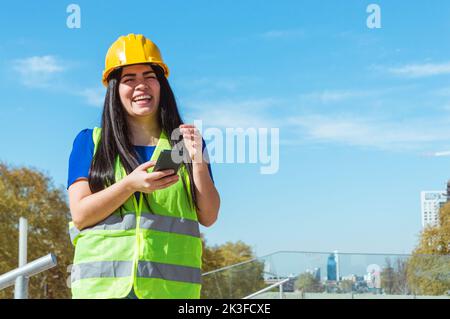 jeune ingénieur vénézuélien latin, portant un casque jaune et un gilet debout à l'extérieur riant à voix haute à cause d'une plaisanterie textée à son téléphone, techn Banque D'Images