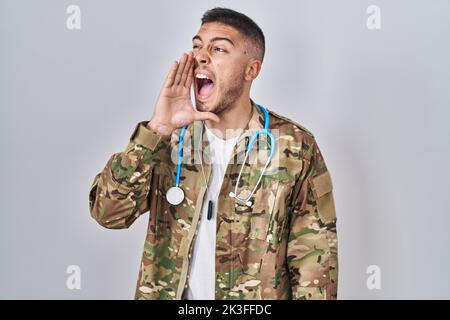 Jeune médecin hispanique portant un uniforme militaire de camouflage sans indice et confondu avec les bras ouverts, aucune idée et visage douteux. Banque D'Images