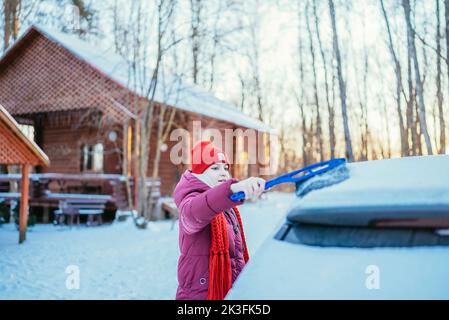 une fille dans un chapeau rouge nettoie une voiture enneigée en hiver par temps froid. Banque D'Images