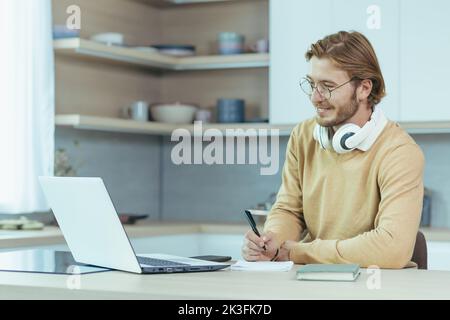 Jeune homme entrepreneur travaillant à la maison sur le travail de papier, assis dans la cuisine à la table, en utilisant un ordinateur portable pour le travail à distance en tant que indépendant. Banque D'Images