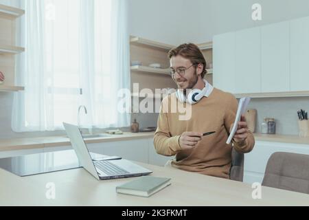 Un jeune enseignant enseigne à distance depuis la maison, un homme travaille dans la cuisine à la maison, utilise un ordinateur portable pour les appels en ligne avec communique avec les élèves en souriant explique le travail montre le bloc-notes. Banque D'Images