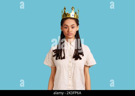 Arrogant femme égoïste avec des dreadlocks noirs regardant égoïsttiquement la caméra, posant avec la couronne sur la tête, prétendant être reine, portant chemise blanche. Studio d'intérieur isolé sur fond bleu. Banque D'Images