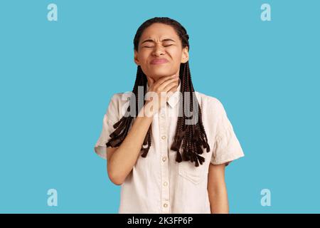 La femme malheureuse avec des dreadlocks a une terrible douleur de gorge, souffre de suffocation, touche le cou, frondes frondes, difficile à avaler, porter une chemise blanche. Studio d'intérieur isolé sur fond bleu. Banque D'Images