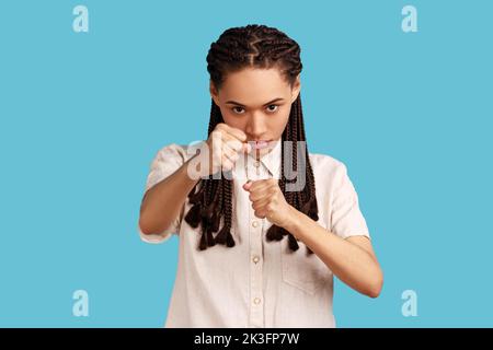 Femme aux dreadlocks noirs qui a un look sérieux et confiant, qui maintient les poings serrés devant elle, se désardu de l'offense et du mauvais traitement. Studio d'intérieur isolé sur fond bleu. Banque D'Images