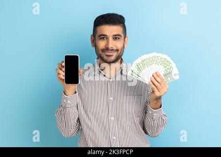 Paiement en ligne. Surpris homme d'affaires avec une barbe tenant un téléphone portable et des billets en euros, choqué par le cashback d'argent, portant une chemise rayée. Studio d'intérieur isolé sur fond bleu. Banque D'Images