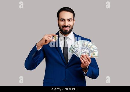 Homme tenant entre les mains et montrant des billets en dollars et une pièce d'or de crypto-monnaie, transférant de l'argent dans des bitcoins numériques, portant le costume de style officiel. Prise de vue en studio isolée sur fond gris. Banque D'Images