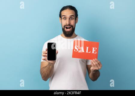 Portrait de l'homme avec la barbe portant le T-shirt blanc largement ouvert les yeux et la bouche tenant la carte de vente et le smartphone, choqué par l'incroyable vente en ligne. Studio d'intérieur isolé sur fond bleu. Banque D'Images