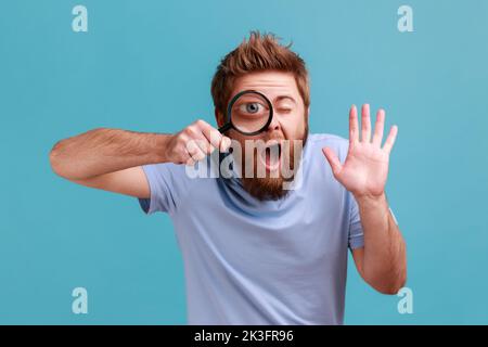Portrait de drôle barbu homme tenant la loupe et regardant la caméra avec grand œil de zoom, visage curieux, la main agissante, disant Bonjour. Studio d'intérieur isolé sur fond bleu. Banque D'Images