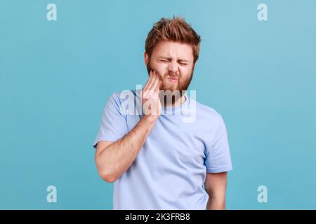 Portrait d'un homme barbu malade malsain qui appuie sur la joue, qui souffre de maux de dents aigus, de maladies parodontales, de cavités ou de douleurs à la mâchoire. Studio d'intérieur isolé sur fond bleu. Banque D'Images