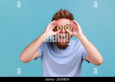 Bitcoin, argent numérique. Portrait d'un homme barbu enjoué et excité couvrant les yeux avec des bitcoins d'or, en étant confiant dans la crypto-monnaie. Studio d'intérieur isolé sur fond bleu. Banque D'Images