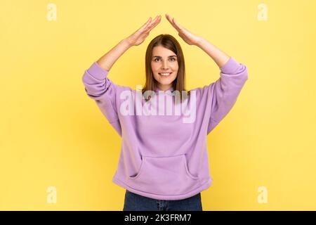 Je suis en sécurité. Portrait d'une femme protégée heureuse levant les mains au-dessus de la tête, faisant un geste de toit, assurance, portant le pull à capuche violet. Studio d'intérieur isolé sur fond jaune. Banque D'Images