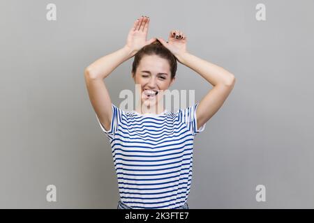 Portrait d'une femme joueur portant un T-shirt rayé faisant des oreilles de lapin drôles avec les mains sur la tête, comportement enfantin, humeur optimiste. Prise de vue en studio isolée sur fond gris. Banque D'Images