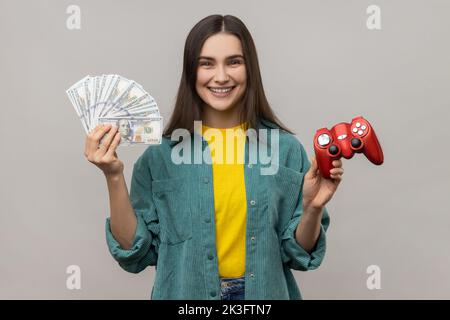 Portrait d'une femme extrêmement heureuse montrant à l'appareil photo rouge joypad et des billets de dollars, jouant et gagnant de l'argent, portant une veste de style décontracté. Prise de vue en studio isolée sur fond gris. Banque D'Images