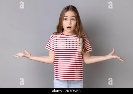 Que voulez-vous ? Portrait d'une petite fille portant un T-shirt rayé debout avec des mains levées et surpris d'expression indignée, demandant quelle raison. Prise de vue en studio isolée sur fond gris. Banque D'Images