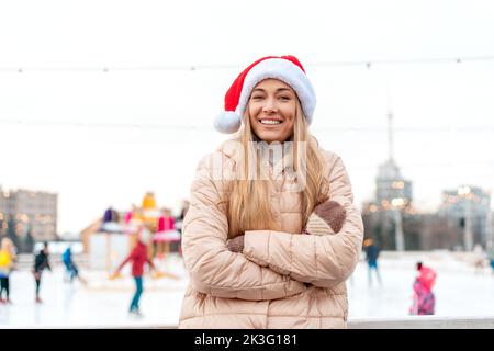 Portrait d'extérieur jeune belle femme à la mode portant chapeau de Santa, posant rue de la ville européenne. Hiver Noël concept de vacances. Patinoire arrière-plan. Vêtements d'hiver caucasiens pour femmes Banque D'Images