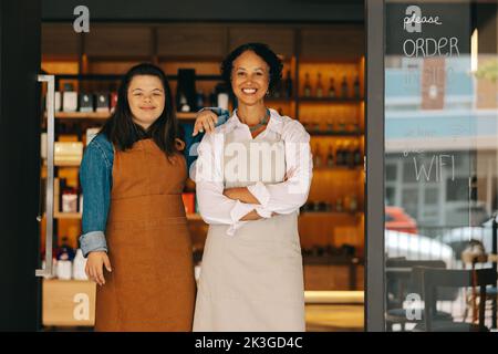 Les employés heureux du magasin sourient à l'appareil photo tout en se tenant à l'entrée de leur magasin d'alimentation. Femme autonome avec le syndrome de Down travaillant dans un loc Banque D'Images