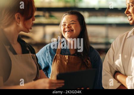 Employé de la vente au détail avec le syndrome de Down souriant heureux tout en se tenant debout dans une réunion de personnel dans une épicerie. Groupe de femmes diverses travaillant ensemble dans un a Banque D'Images