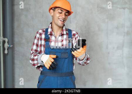 homme souriant travailleur ou constructeur en casque jaune et combinaison avec smartphone sur fond de mur gris. profession, construction et bâtiment co Banque D'Images