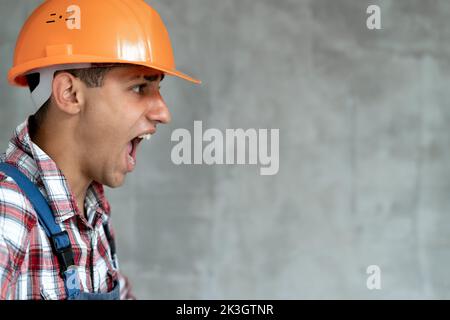 Jeune homme de construction portant un uniforme de construction et un casque de sécurité sur fond gris isolé criant fou et fou avec une expression agressive. FRU Banque D'Images