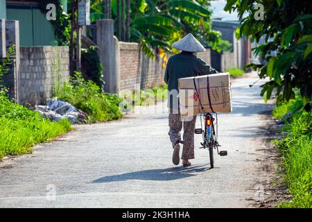 Homme vietnamien avec chapeau de bambou poussant vélo à travers le village rural de montagne Hoang Mai, Hai Phong, Vietnam Banque D'Images