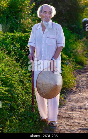 Vieil homme vietnamien avec cheveux blancs, barbe, et chapeau de bambou debout au bord de la route, rural Hai Phong, Vietnam Banque D'Images