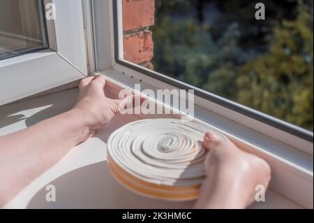 une femme colle un ruban de caoutchouc d'étanchéité sur une fenêtre dans un salon. Banque D'Images