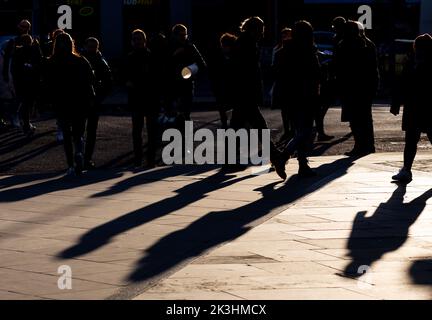 Une foule de personnes en silhouette traversant la route créant de longues ombres.le soleil rétro-éclairé donne une atmosphère dramatique à la photo Banque D'Images