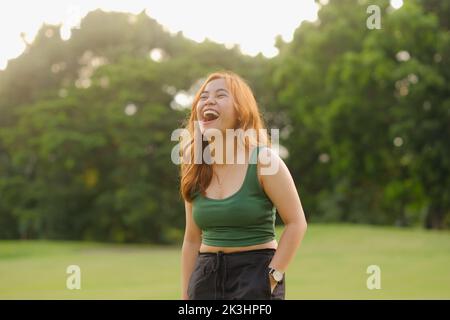 Jeune femme asiatique avec de longs cheveux ondulés de caramel et le haut de réservoir vert howls avec rire. Cadre extérieur avec arbres et herbe. Lumière naturelle au coucher du soleil. Banque D'Images