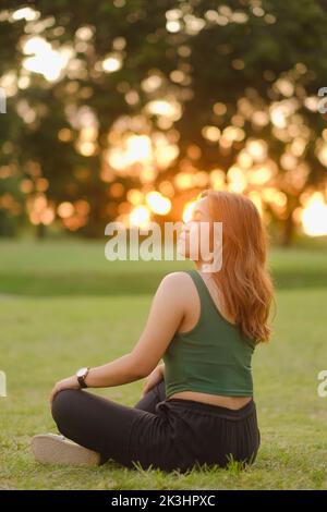 La jeune femme asiatique aux longs cheveux ondulés, au débardeur vert et au pantalon noir est assise à pieds croisés sur l'herbe et au coucher du soleil avec les yeux fermés. Banque D'Images