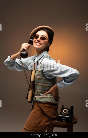 bonne jeune femme en béret et lunettes de soleil tenant téléphone vintage sur marron, image de stock Banque D'Images