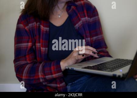 Femme courte non reconnaissable en chemise à carreaux avec ordinateur portable sur les jambes. Travailleur indépendant et travailleur indépendant travaillant à domicile Banque D'Images