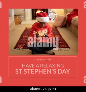 Composition du texte du jour de St stephen et petit garçon biracial en chapeau de santa à noël par cheminée Banque D'Images