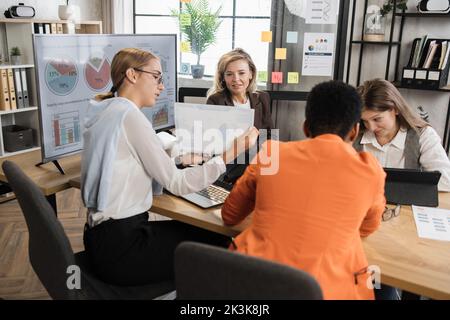 Quatre femmes économistes multiethniques siégeant au bureau avec divers gadgets et analysant la croissance financière de l'économie mondiale. Femmes partenaires ayant une réunion d'affaires. Banque D'Images
