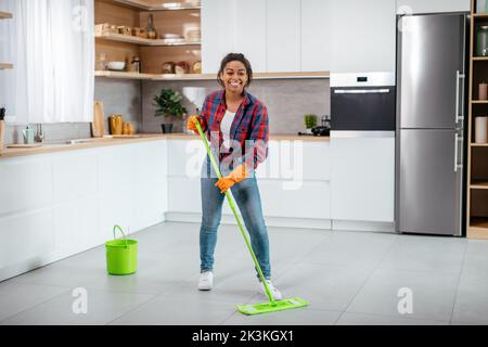 Bonne jeune femme noire en gants de caoutchouc lave le sol avec une vadrouille dans un intérieur minimaliste de cuisine, sur toute la longueur Banque D'Images