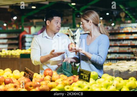 Un jeune couple international, une femme et un homme asiatique, choisissent des fruits dans un supermarché, décidant quelles pommes acheter. Ils sont dans le département des fruits et légumes avec un sac en plastique. Banque D'Images