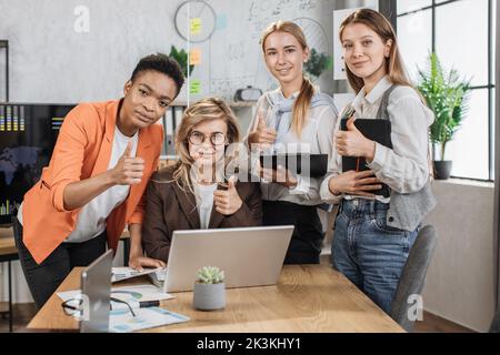 Belle patron de femme âgée assise à la table au bureau et à l'écoute de ses divers collègues montrant le pouce, se tenant autour et discutant des résultats d'un travail ou d'une stratégie conjoints. Banque D'Images