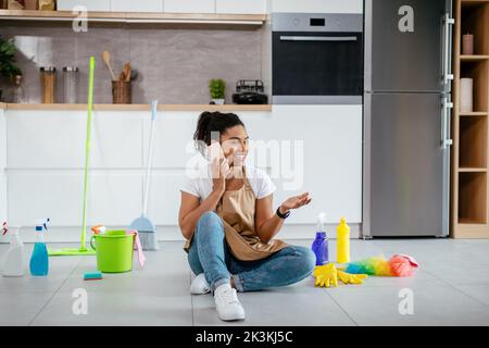 Une jeune femme noire heureuse est assise sur le sol avec des articles de nettoyage par téléphone dans l'intérieur moderne de la cuisine Banque D'Images