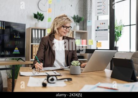 Portrait d'une charmante femme âgée dans un costume élégant et formel, avec des notes et un ordinateur portable moderne, assis au bureau. Plutôt un employé de l'entreprise Banque D'Images