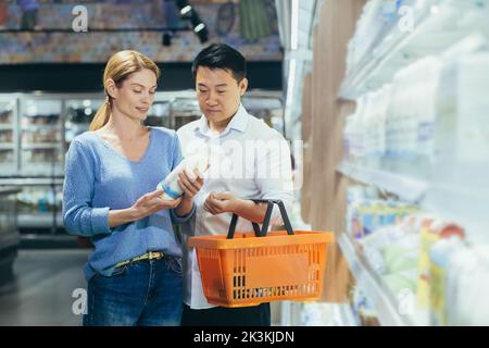 Shopping en famille. Jeune couple interracial, asiatique homme et femme choisissant le lait dans le supermarché. Ils tiennent un panier et une bouteille de lait dans leurs mains, lire l'étiquette, se tenant près du réfrigérateur. Banque D'Images