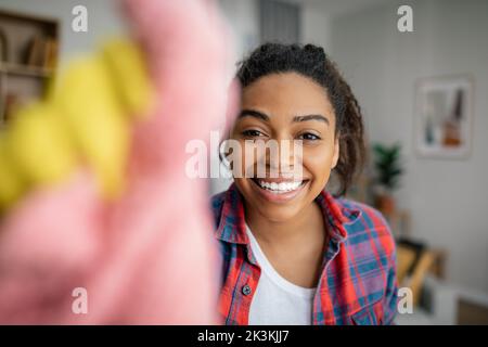 Gaie jeune femme noire dans des gants en caoutchouc lave le miroir avec un chiffon, aime le nettoyage dans l'intérieur de la salle de séjour Banque D'Images