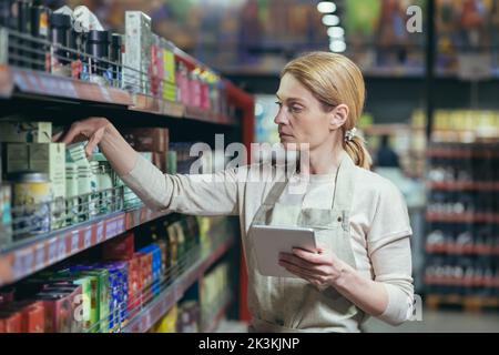 Une belle jeune femme employée d'un supermarché, stocker dans un uniforme de travail affiche les marchandises sur l'étagère, compte, écrit dans un carnet. Banque D'Images
